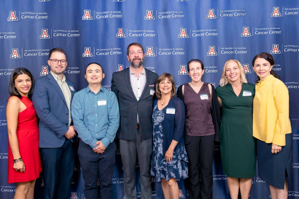 Eight members of the University of Arizona Cancer Center stand side-by-side smiling in front of a Cancer Center backdrop. 