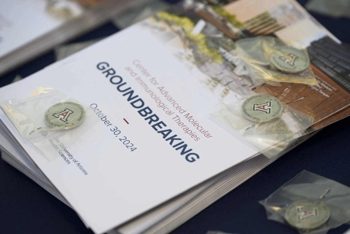 A stack of event programs with small, silver lapel pins scattered around it sits on a table. The program cover reads “Groundbreaking.” 