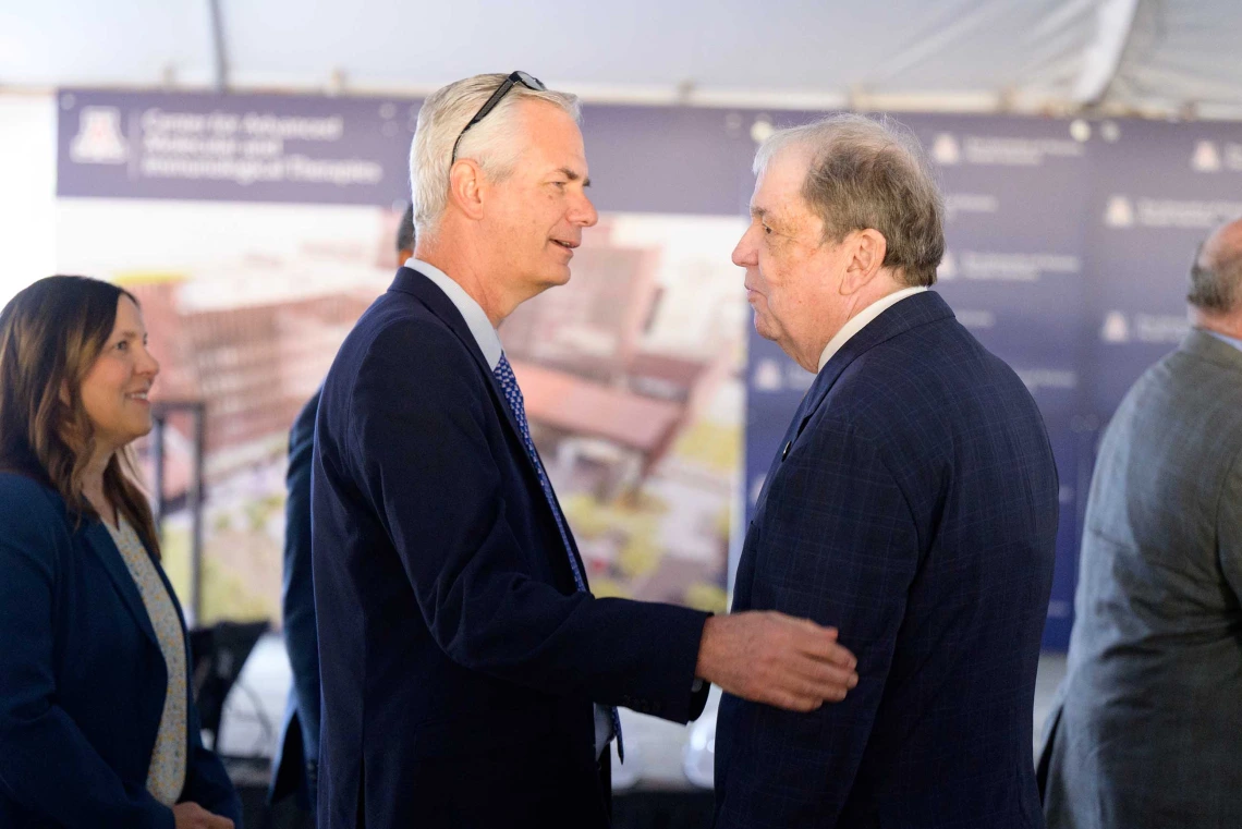 Two men in dark blazers face each other as they talk. One has his hand on the other’s elbow.  