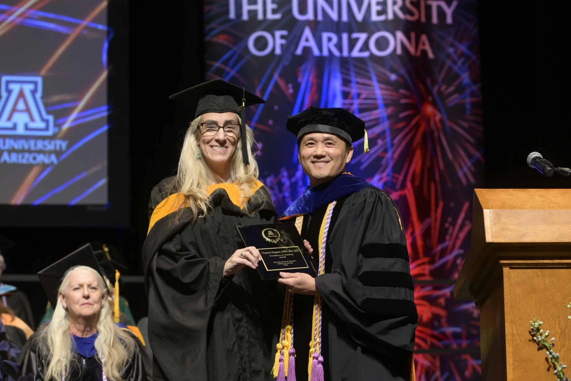 Two University of Arizona College of Nursing faculty members dressed in graduation regalia stand together on a stage, smiling and holding a plaque.  