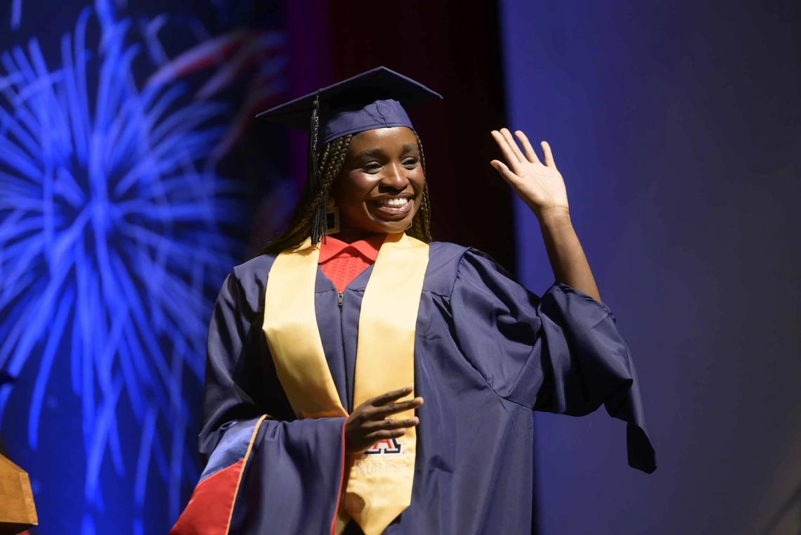 A woman in graduation regalia waves and smiles as she walks across a stage. 