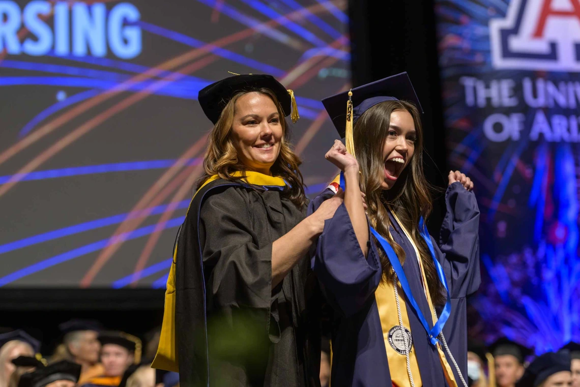 A young women in a graduation cap and gown pumps her arms in the air and smiles as a professor puts a ribbon around her neck