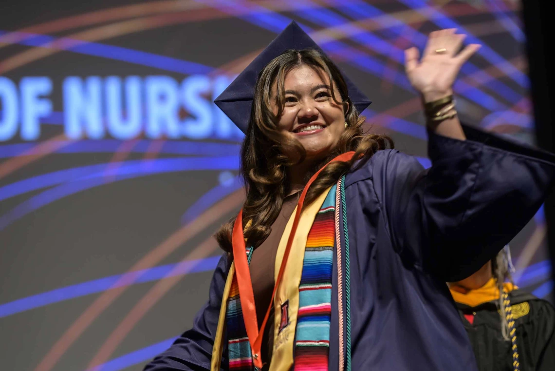 A young woman wearing graduation regalia smiles and waves as she walks across a stage. 