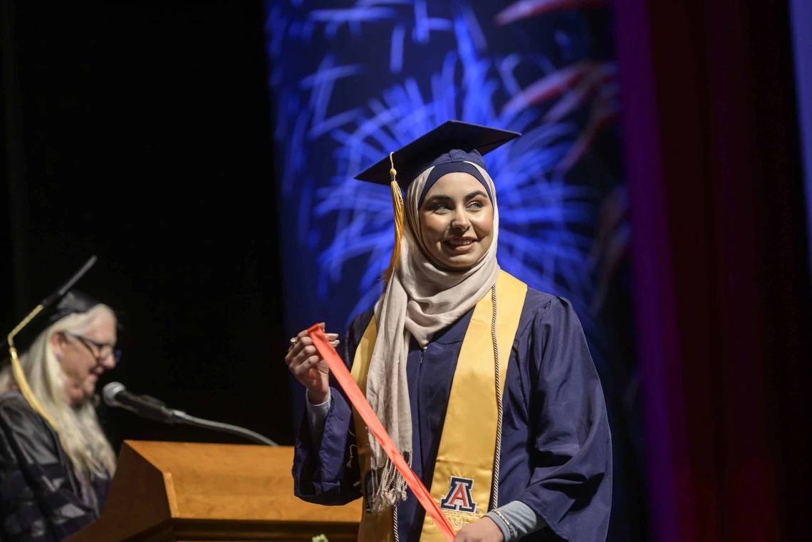 A woman wearing a hijab and traditional graduation regalia smiles as she walks across a stage. 