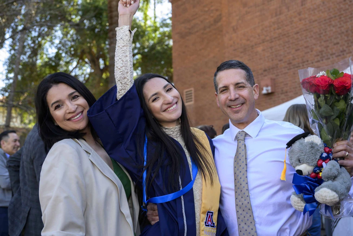 A young woman wearing a graduation gown raises her arm in celebration as she stands between her smiling parents. 