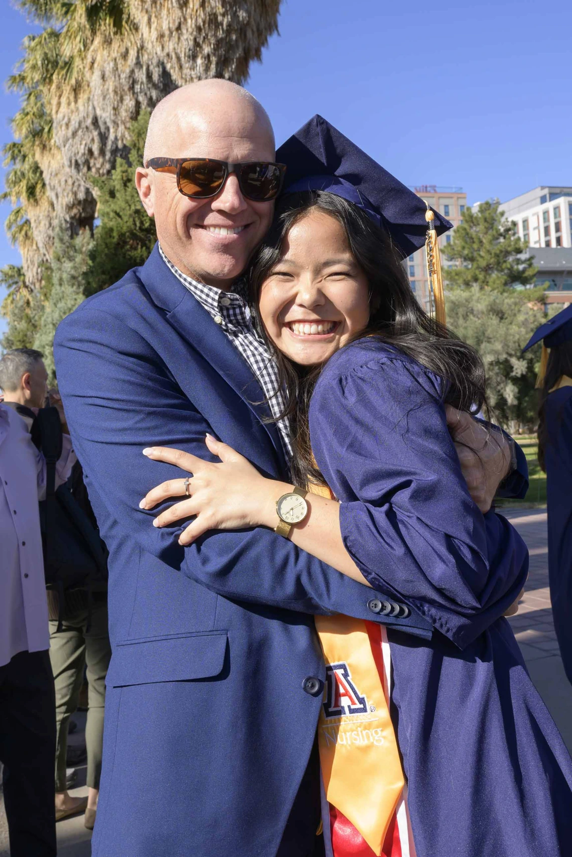 A young women dressed in a graduation cap and gown smiles as she hugs her smiling father.