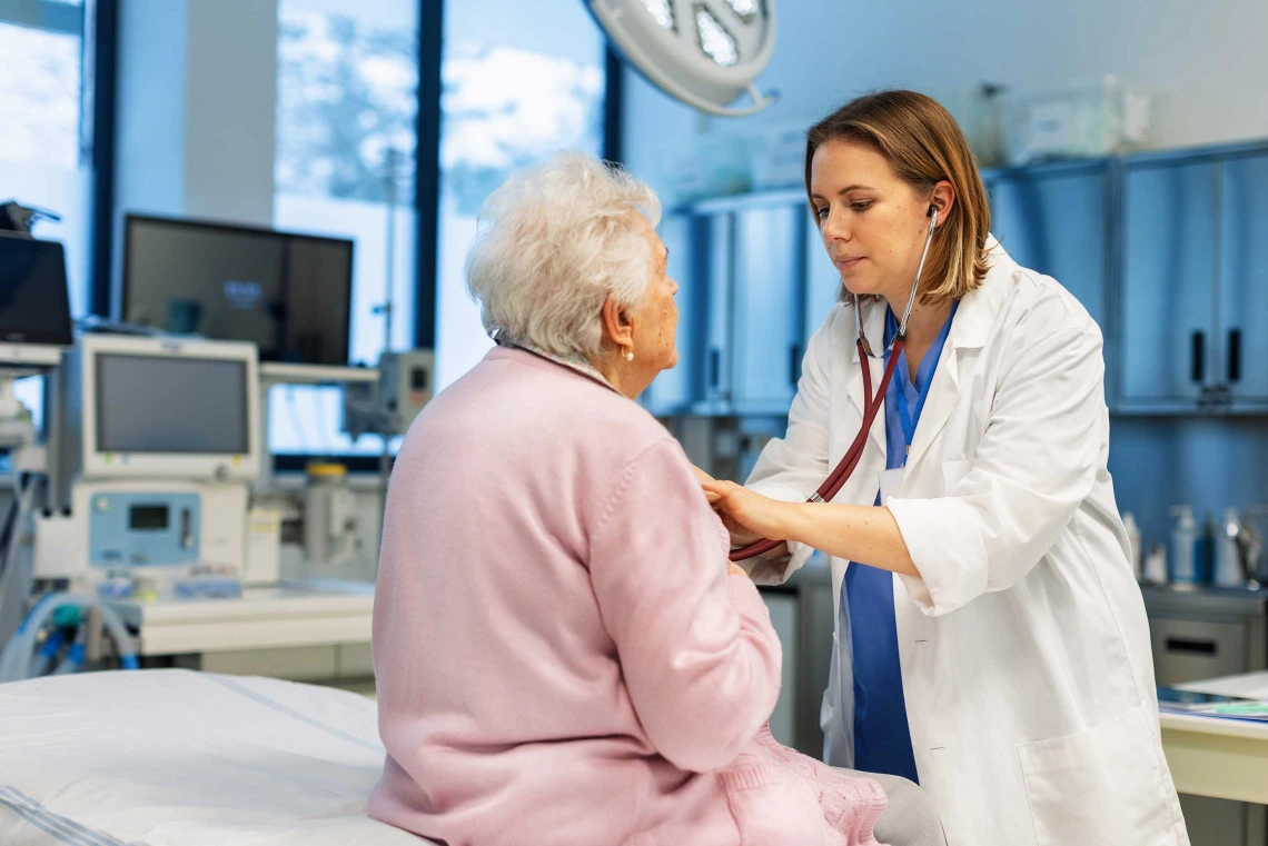 A doctor examines a patient wearing a pink robe sitting on the edge of a bed