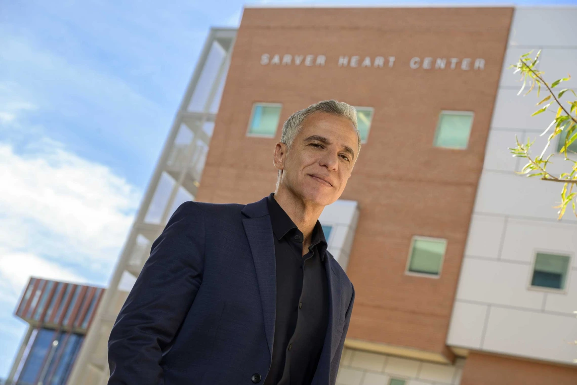 A man in a dark suit coat and black shirt stands outside a building that has Sarver Heart Center signage.