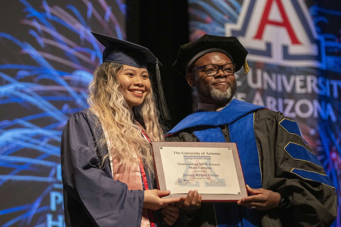 A student and a professor wearing graduation regalia stand next to each other, smiling and holding a certificate. 