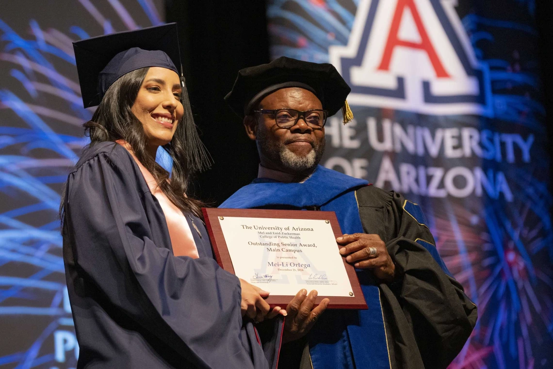 A student and a professor wearing graduation regalia stand next to each other, smiling and holding a certificate.