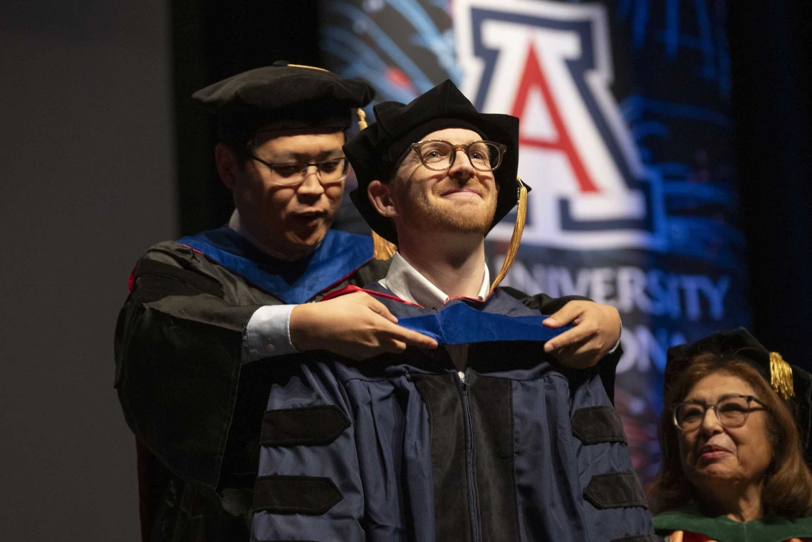 A professor places a sash on the shoulders of a graduate. Both are wearing caps and gowns. 