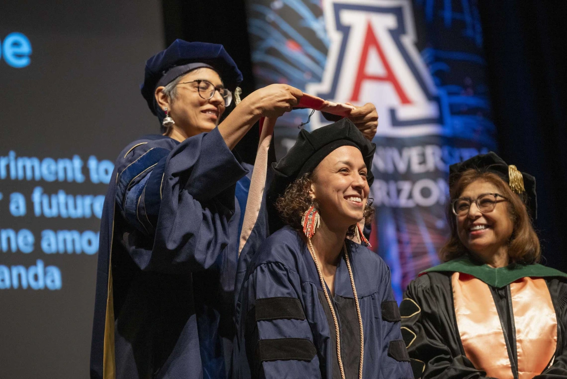 A professor places a sash on the shoulders of a graduate. Both are wearing caps and gowns.