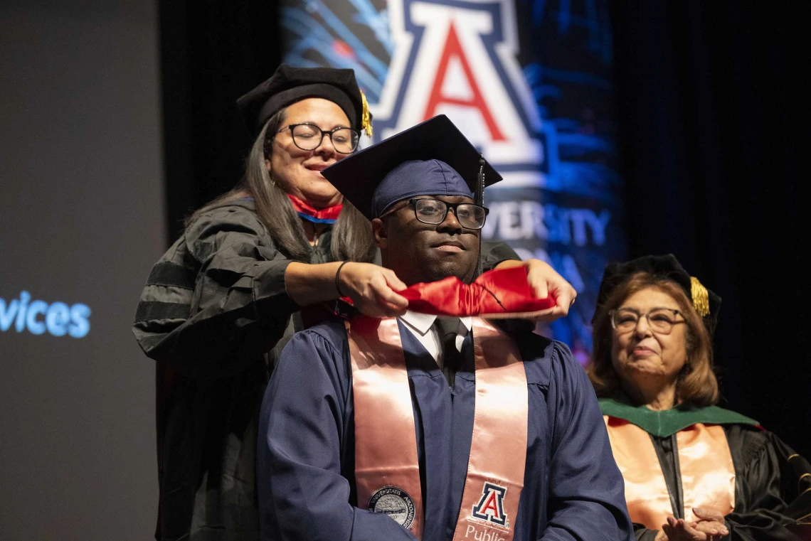 A professor places a sash on the shoulders of a graduate. Both are wearing caps and gowns.