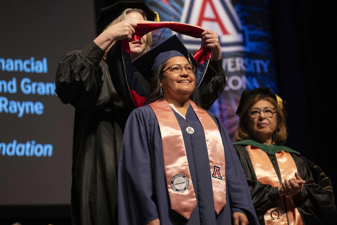 A professor places a sash on the shoulders of a graduate. Both are wearing caps and gowns.