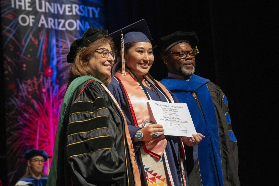 Professors stand on each side of a graduate, who is holding a certificate. All are wearing graduation caps and gowns. 