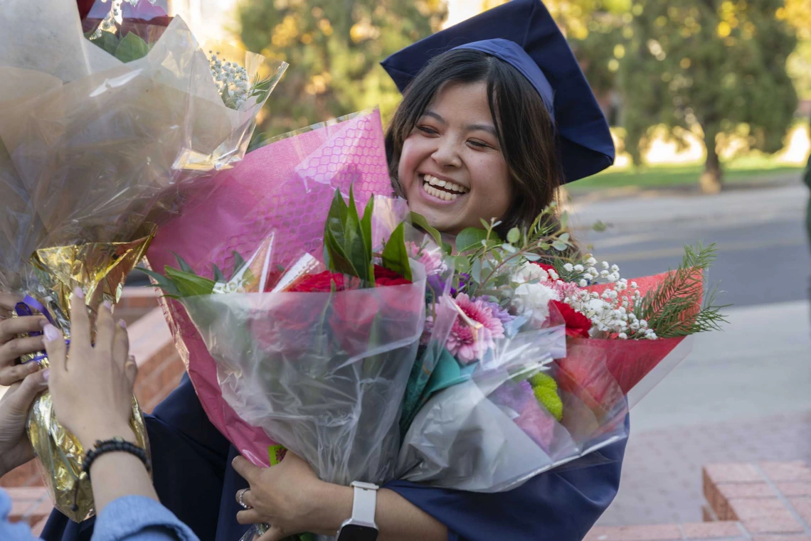 A smiling graduate stands outside with her arms full of flowers. 