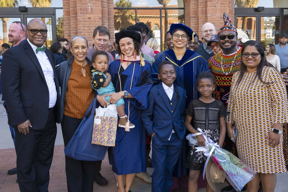 A graduate wearing a cap and gown holds a child while standing with family and friends outside after her graduation ceremony.