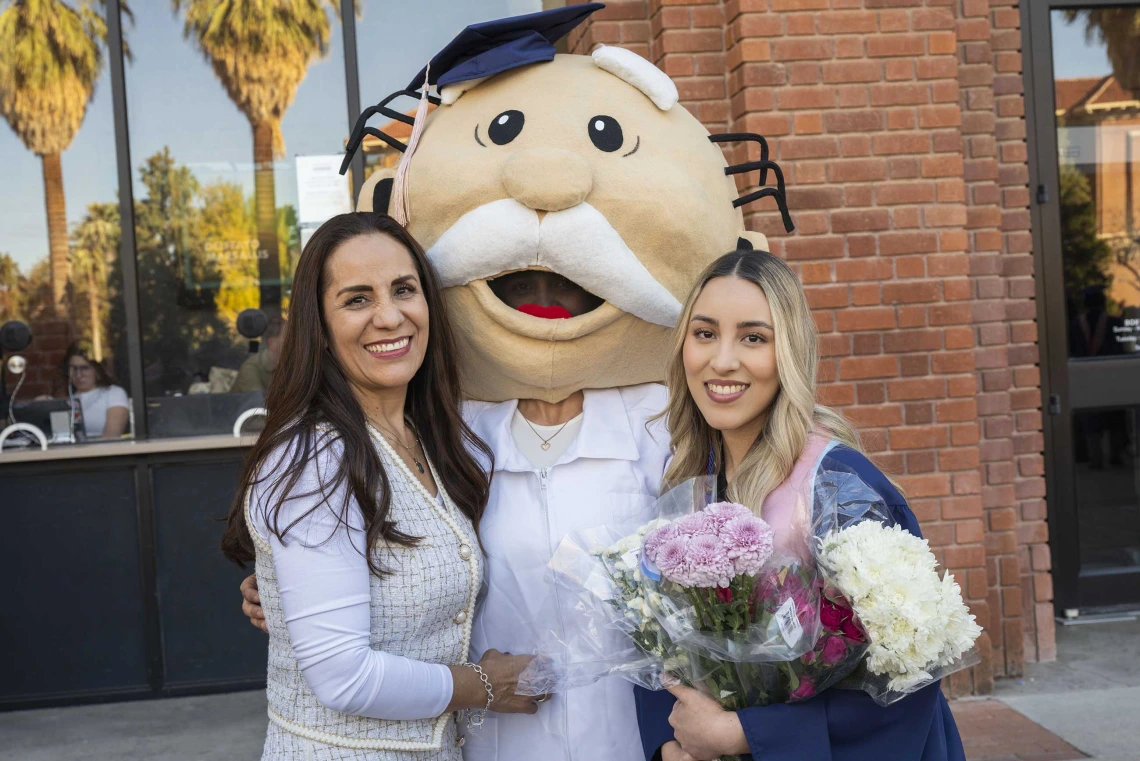 A graduate wearing a cap and gown smiles as she holds flowers while standing next to her mom and her sister, who is wearing a large mascot head adorned with a graduation cap. 