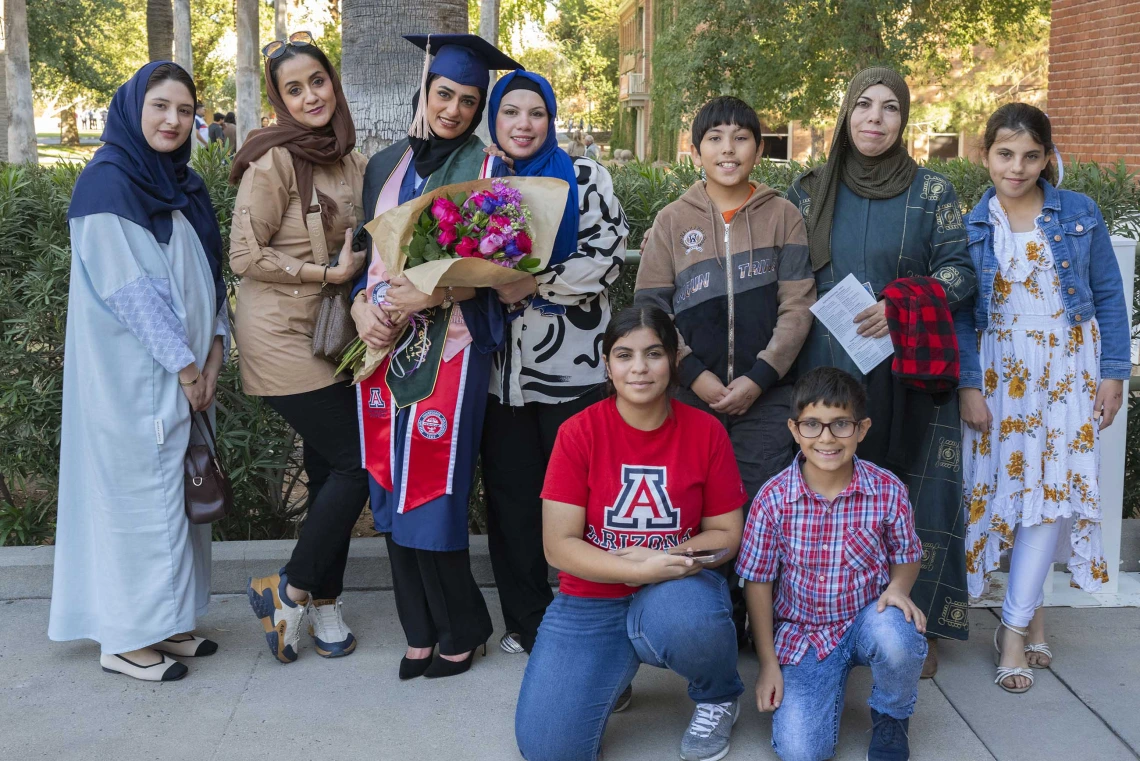 A graduate wearing a cap and gown and holding flowers stands with eight members of her family outside after her graduation ceremony. 