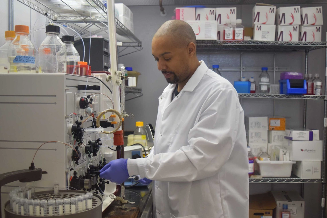 University of Arizona Health Sciences researcher Michael Johnson, dressed in a lab coat and wearing purple gloves, turns knobs on a piece of equipment.