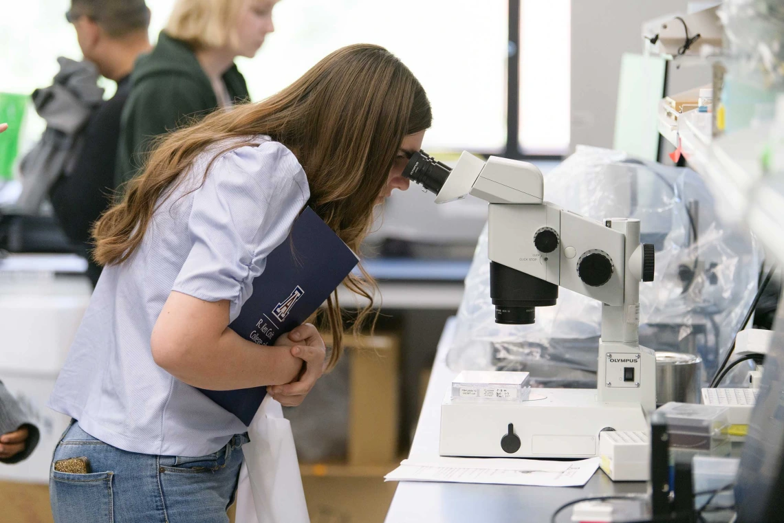 A student holds a folder to her chest while looking into a microscope.