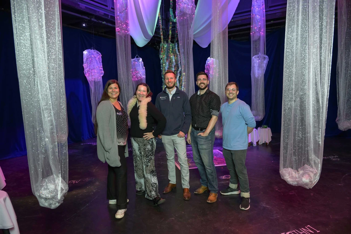 Five people post in the middle of the Hearing the Invisible immersive installation surrounded by columns of mesh fabric hanging from the ceiling