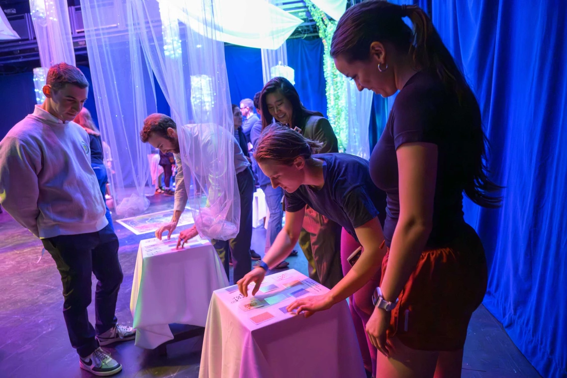 Three people standing around a small table watch as one person plays a card-marching memory game