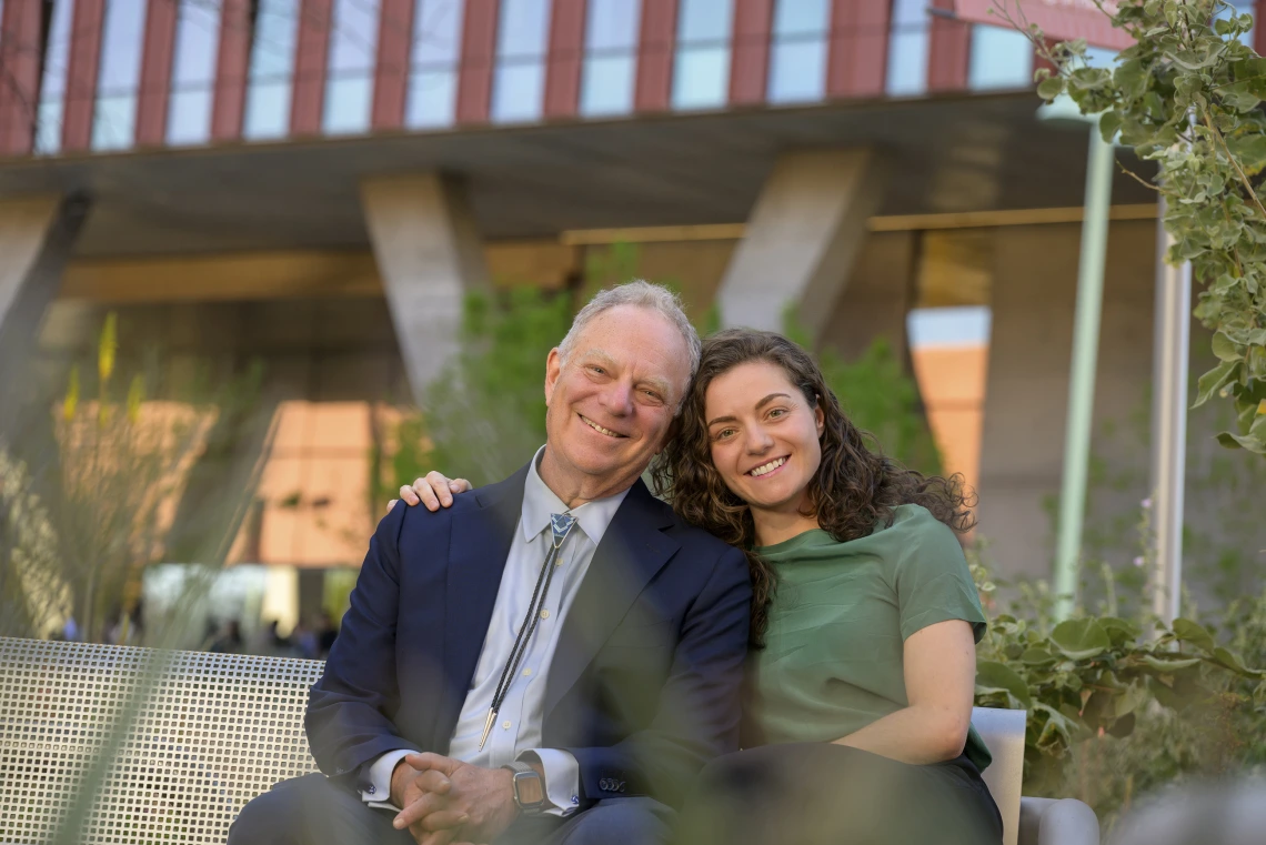 Geoffrey Rubin, MD, MBA, sits next to his daughter Elka Rubin on a bench outside. Their heads are leaned together, touching, and Elka’s arm is around her dad.