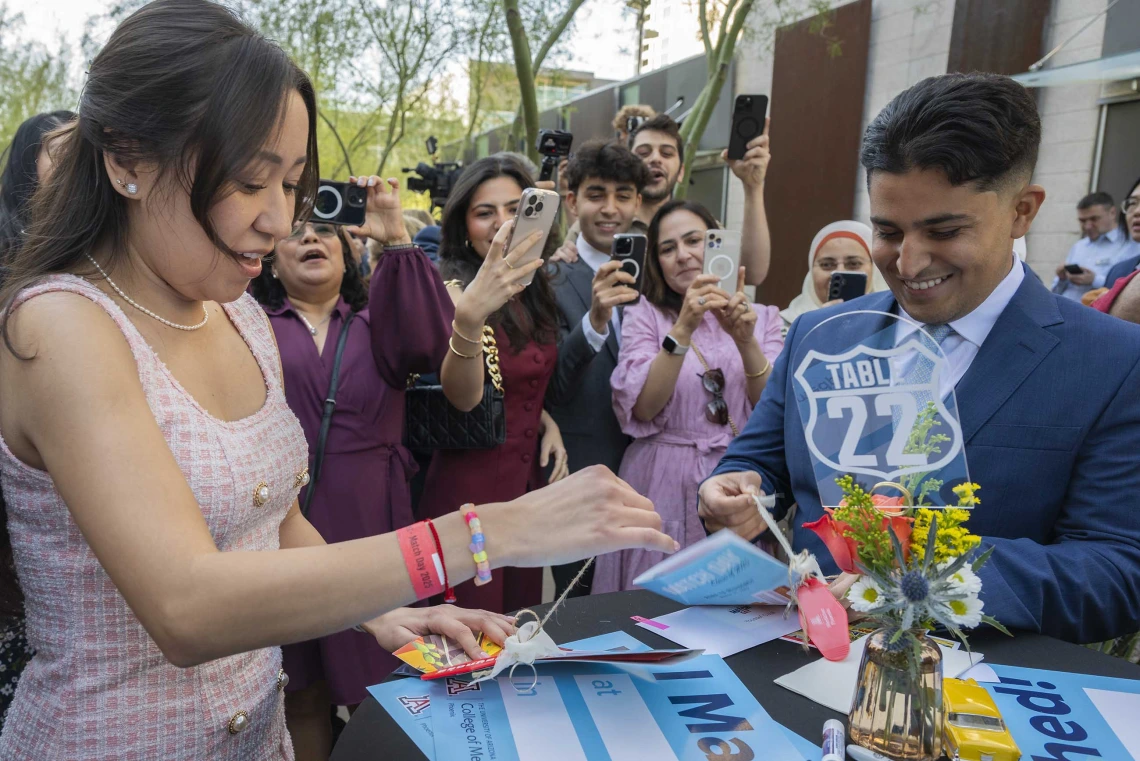 Two well-dressed medical students open envelopes while several family members stand around them with phones in the air taking photos and videos.  