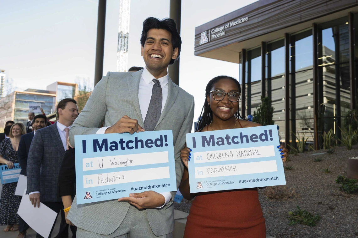 A young man and woman stand next to each other smiling and holding signs that read, “I matched!” in an outdoor setting. 