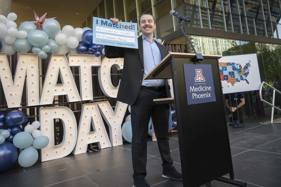 A medical student in a suit stands in front of a lectern holding up a small sign and smiling. 