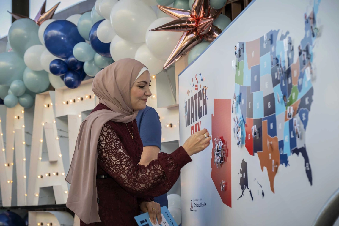 A woman wearing a hijab faces a map of the United States as she prepares to put a pin on the state of Arizona. 