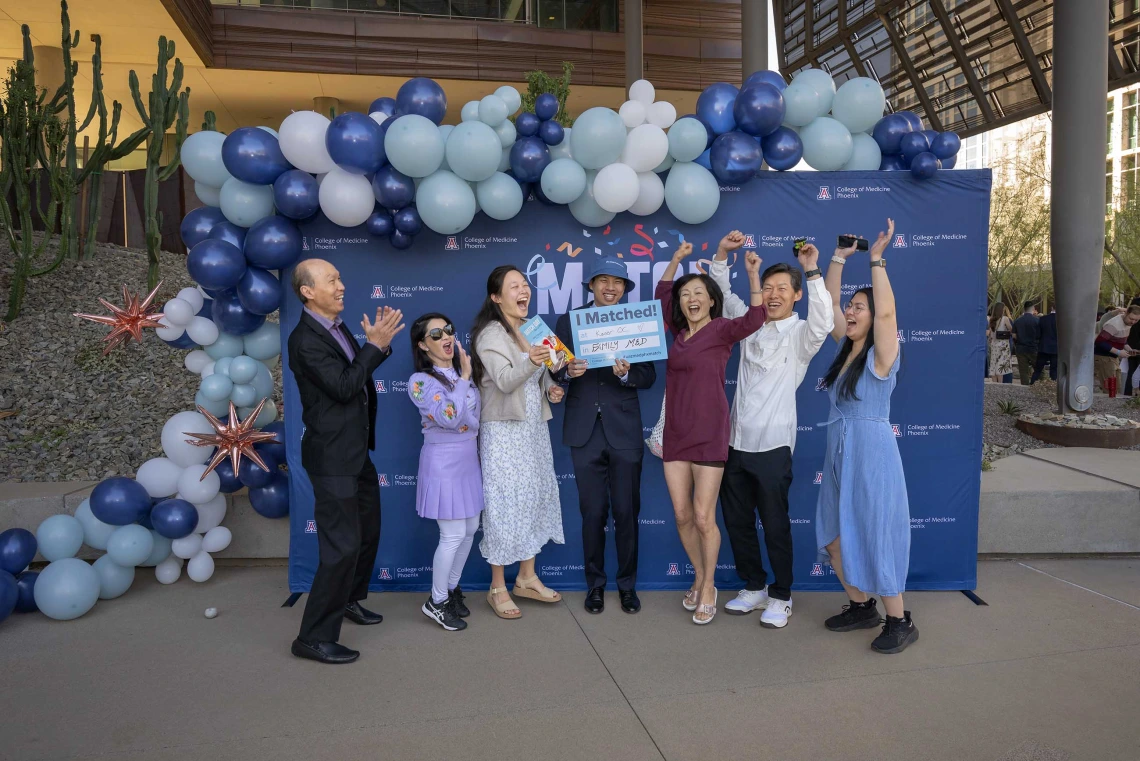 A group of seven cheering people stand in front of a blue background with balloons. 