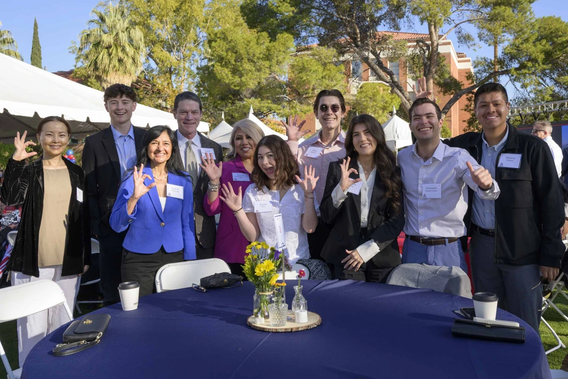 A group of about 10 students and faculty members stand outside behind a table, smiling and making the Arizona Wildcats hand sign. 