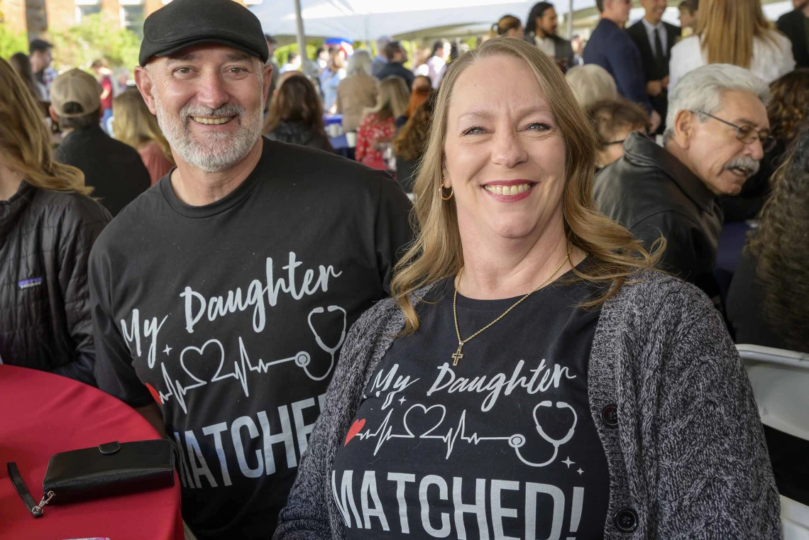 A man and woman sit at a table smiling with shirts that read, “My daughter matched!”