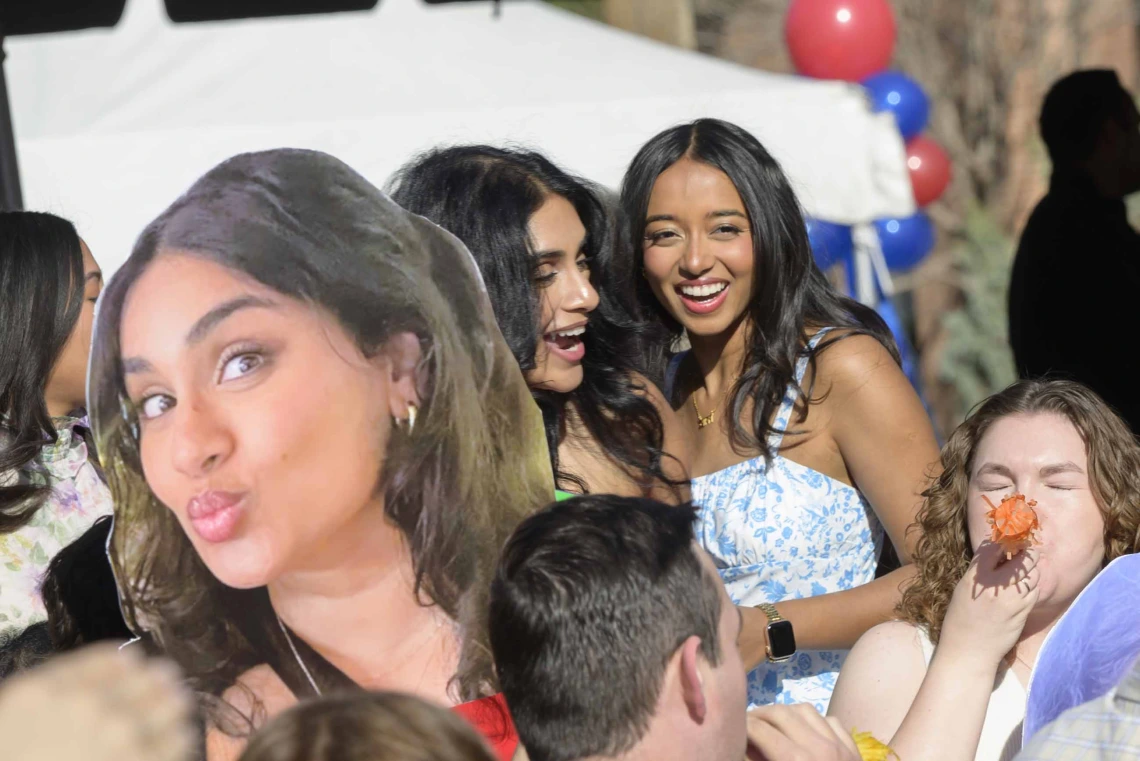 A large photo cutout of a woman’s face is held up in front of the same woman as she smiles and talks with another woman. 