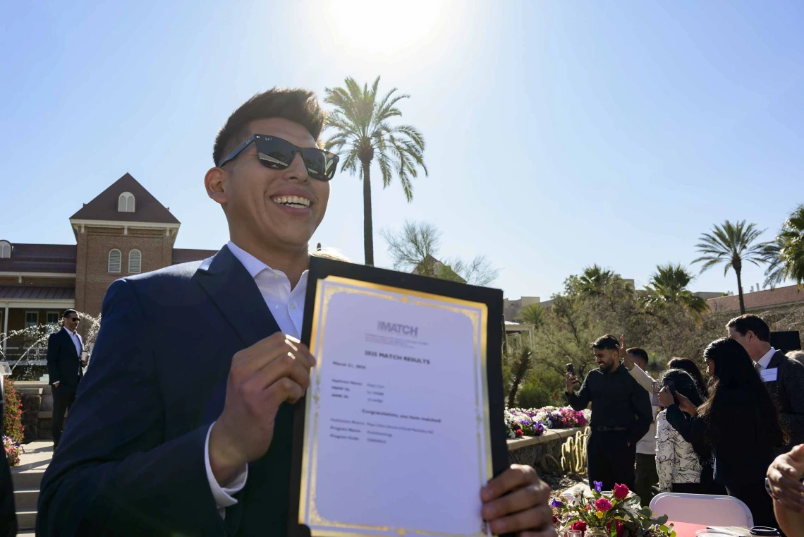 A young man in a suit and sunglasses smiles as he holds up a letter in an outdoor setting. 