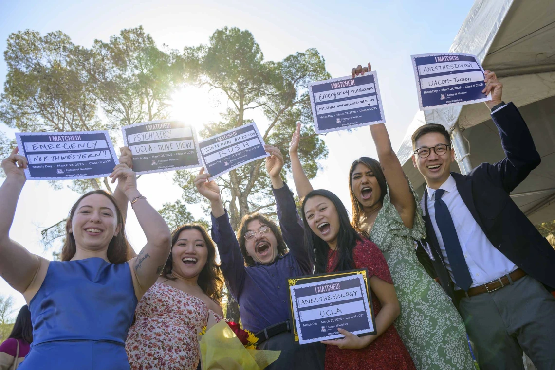 Six medical students smile and cheer as they hold up signs that read, “I matched!” in an outdoor setting. 