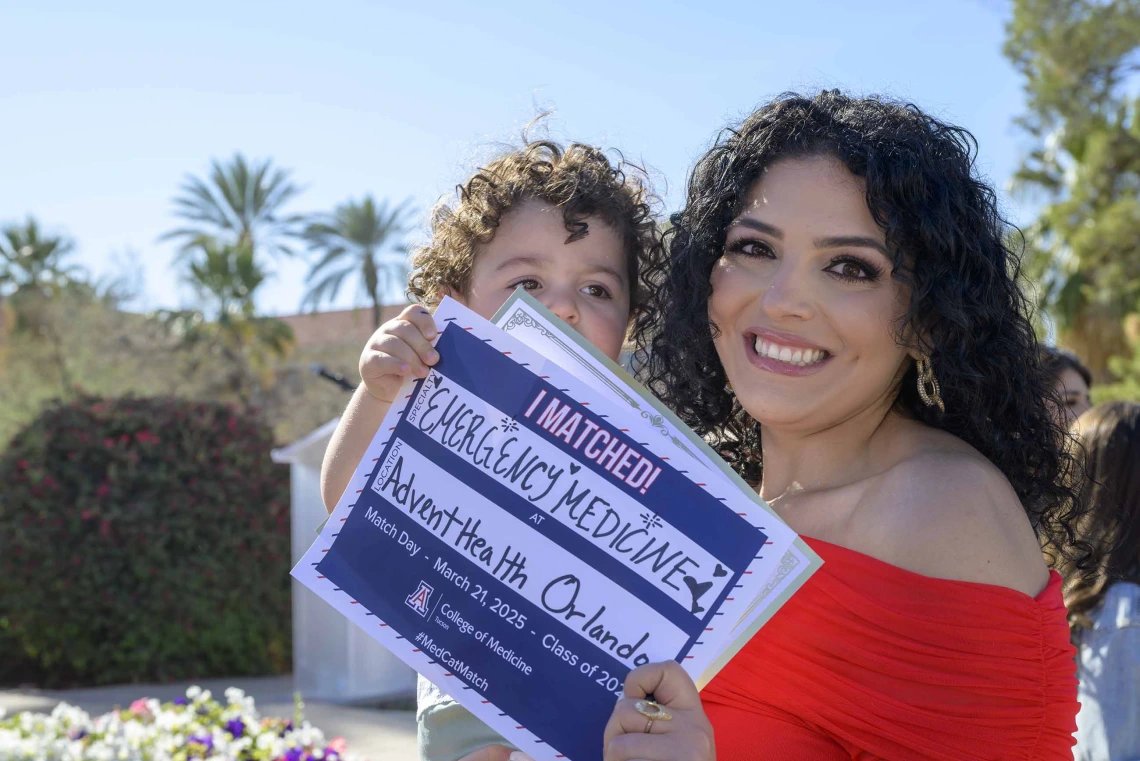 A woman in a red dress smiles as she holds a young boy and a sign that reads, “I matched!”.