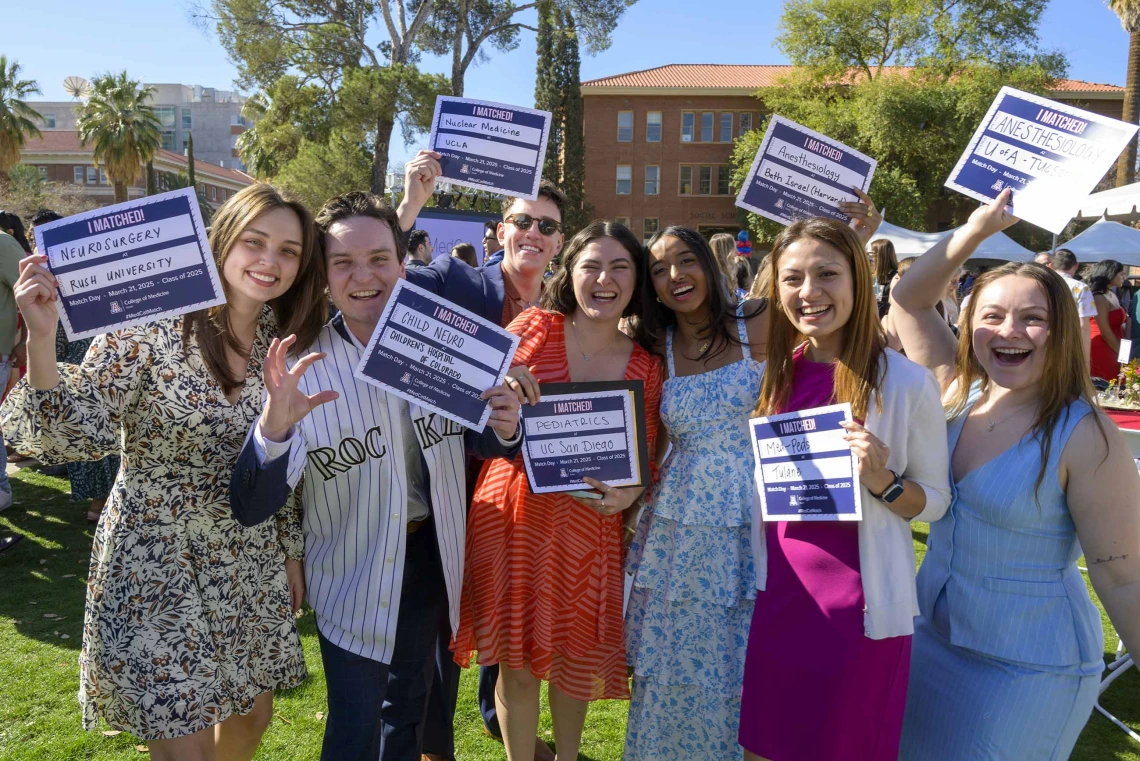 Seven well-dressed medical students stand outside holding small signs that announce where they will be going for their residencies. All are happy and smiling. 