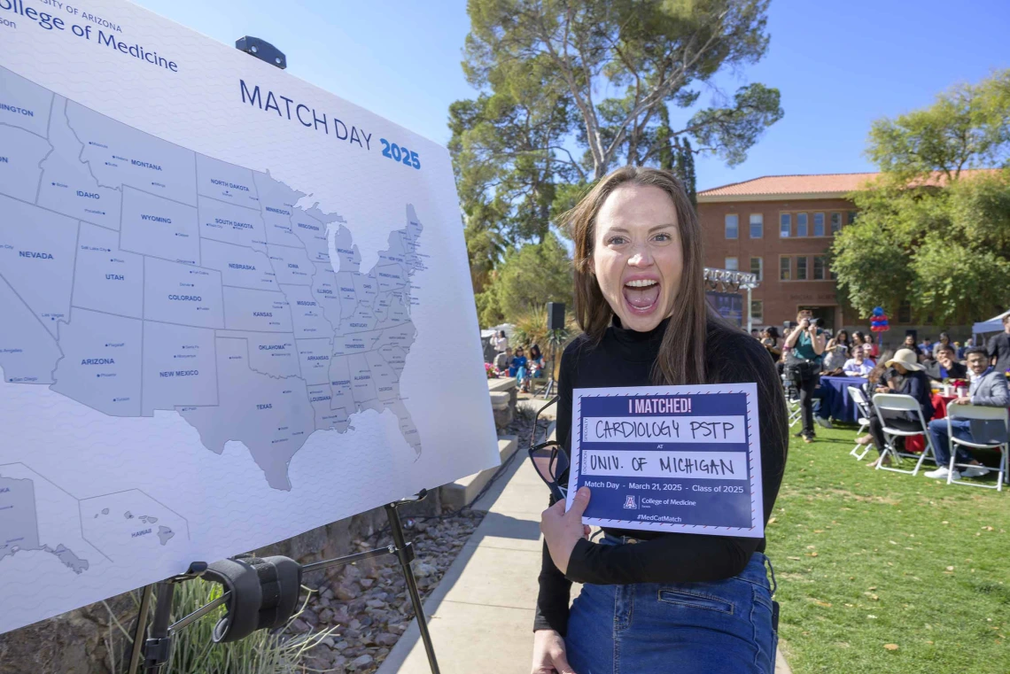 A woman holds up a piece of paper with writing on it noting that she will conduct her medical residency at the University of Michigan Hospital. She has an excited look on her face, with her mouth wide open. 