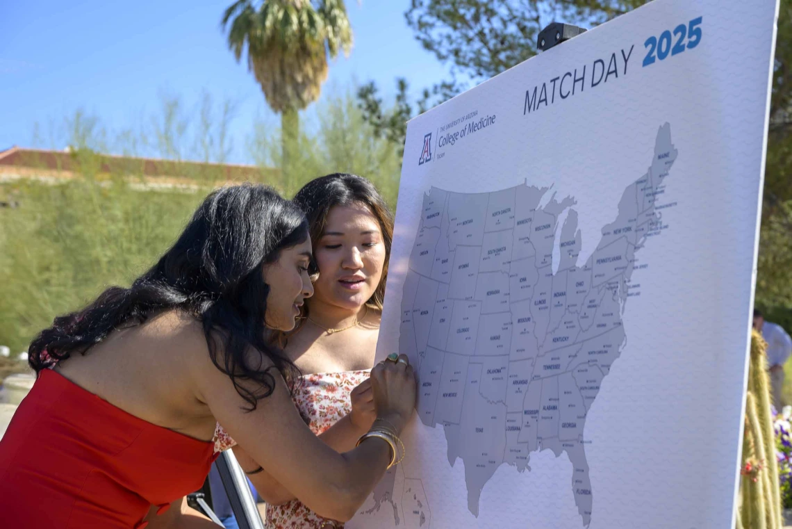 A young woman in a red dress puts a pin in a map of the United States while another young woman looks on. 