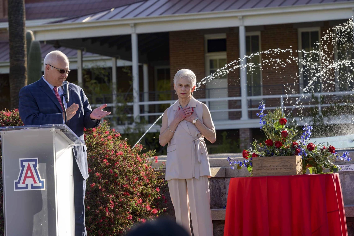 A man in a suit stands behind a lectern clapping as a woman stands nearby with her hands clasped to her heart. They are in an outdoor setting next to a table with flowers and a fountain in the background. 