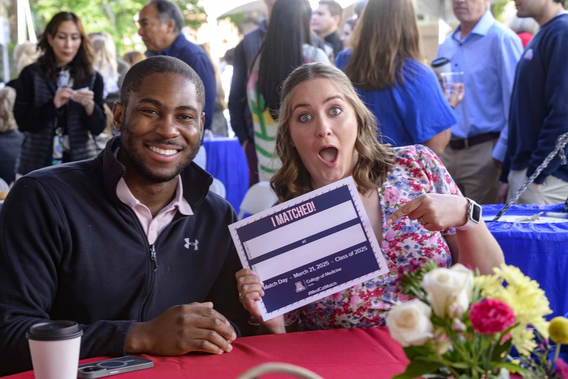 A man and woman sit at a table with flowers as the man smiles and the woman holds up a piece of paper with empty boxes on it. She has a surprised face. 