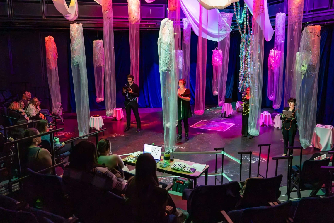 Five singers stand several feet apart on a stage with columns of translucent pink and blue mesh hanging from the ceiling between them