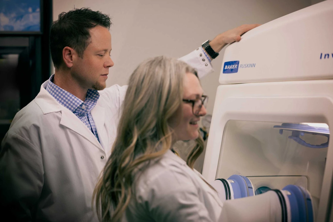 A researcher oversees a student using a glove box in a laboratory at the University of Arizona Cancer Center.