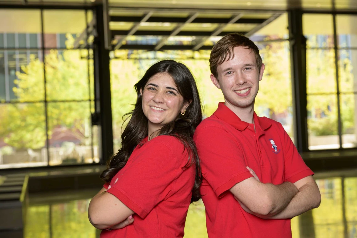 Two students standing back to back with their arms crossed in the lobby of the Health Sciences Innovation Building.