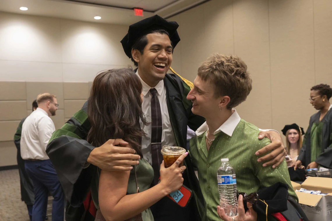 A tall young man wearing a graduation cap and gown smiles as he hugs two friends. 