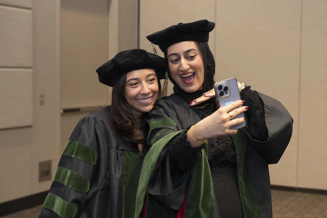 Two medical students in graduation caps and gowns smile as they take a selfie. 