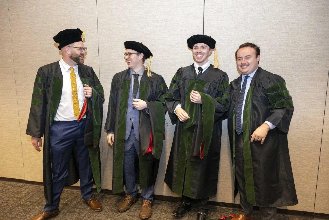Four young men in graduation caps and gowns stand around, laughing. One of them has turned up his cap as a joke. 